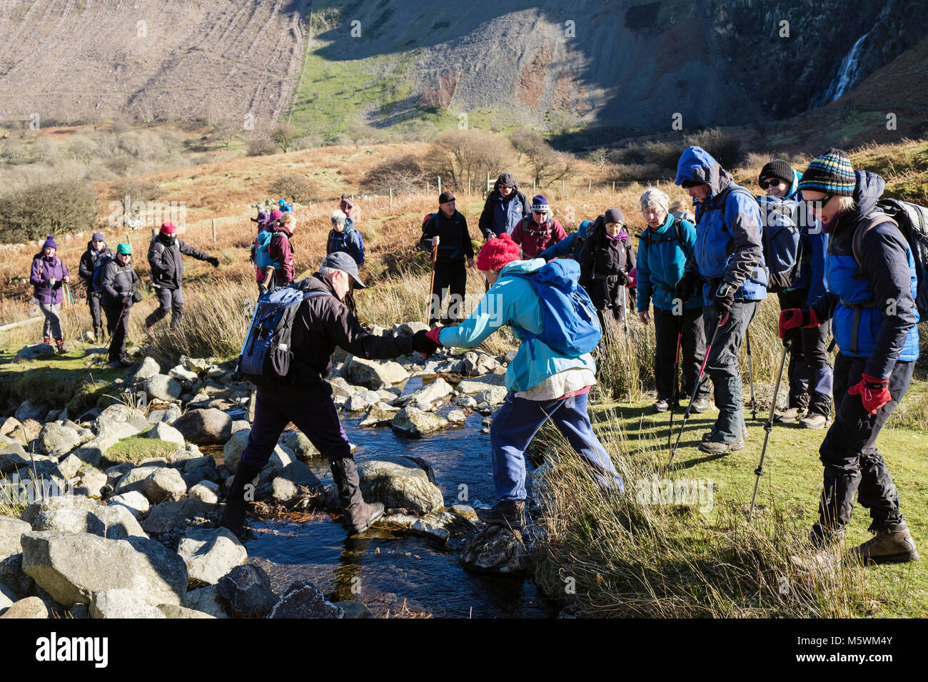 Ramblers group of hikers crossing a stream with man helping someone to cross safely. Abergwyngregyn, Gwynedd, Wales, UK, Britain Stock Photo