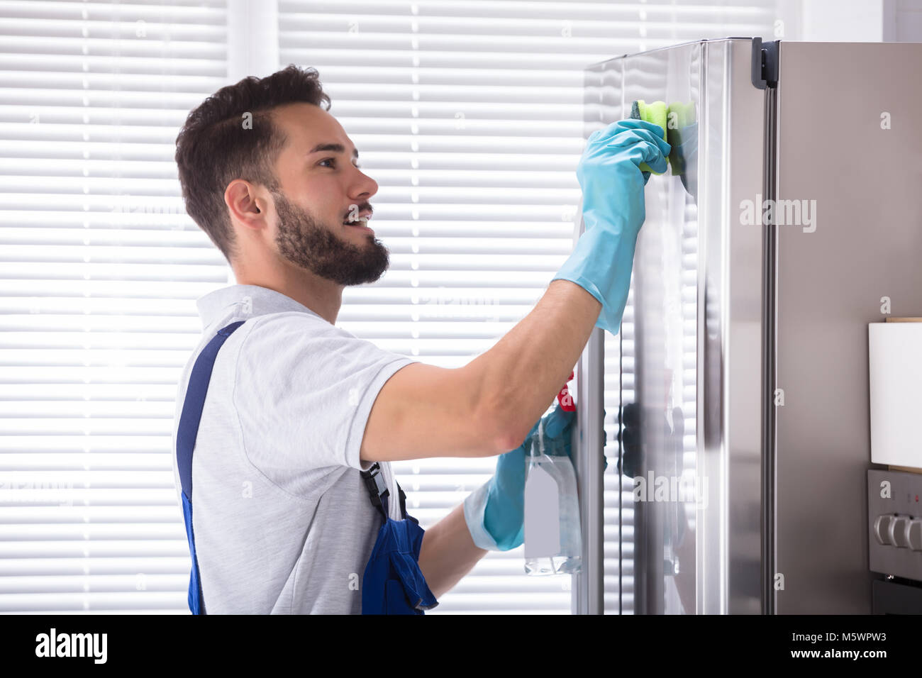 Side View Of A Young Male Janitor Cleaning Refrigerator In Kitchen Stock Photo