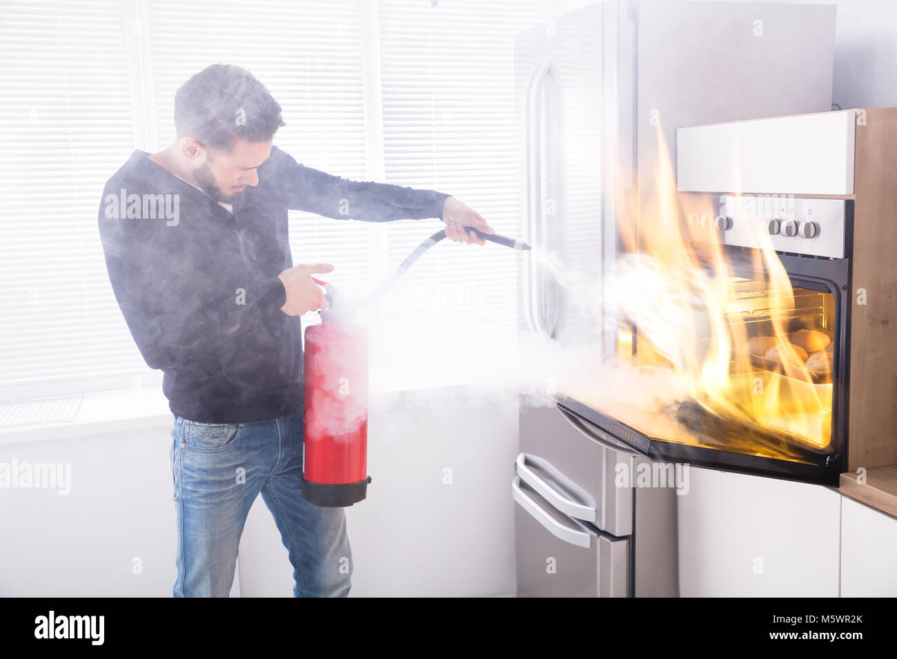 Young Man Using Red Fire Extinguisher To Stop Fire Coming From Oven In