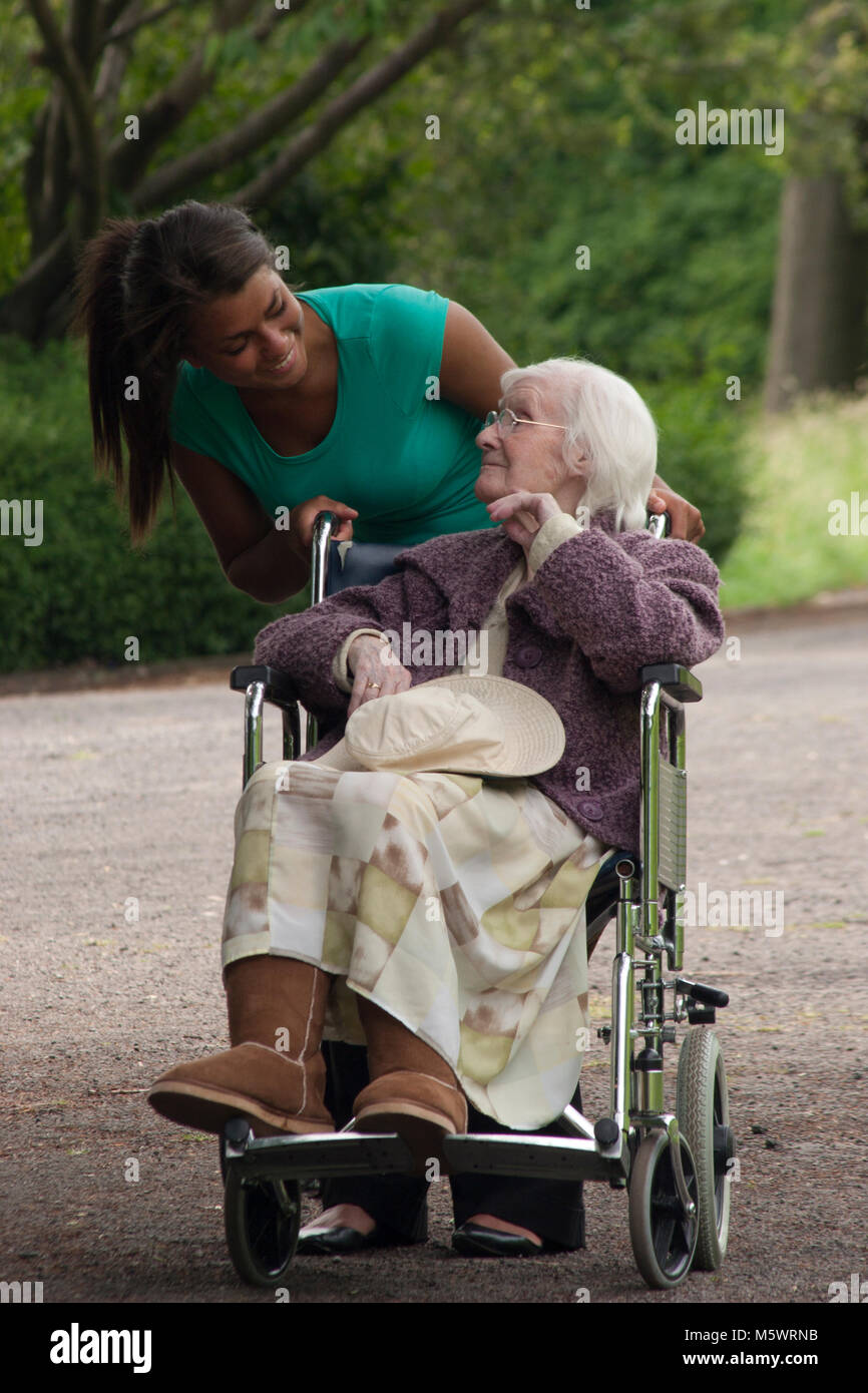 Multicultural young woman carer support worker pushing geriatric lady in wheelchair in the park Stock Photo