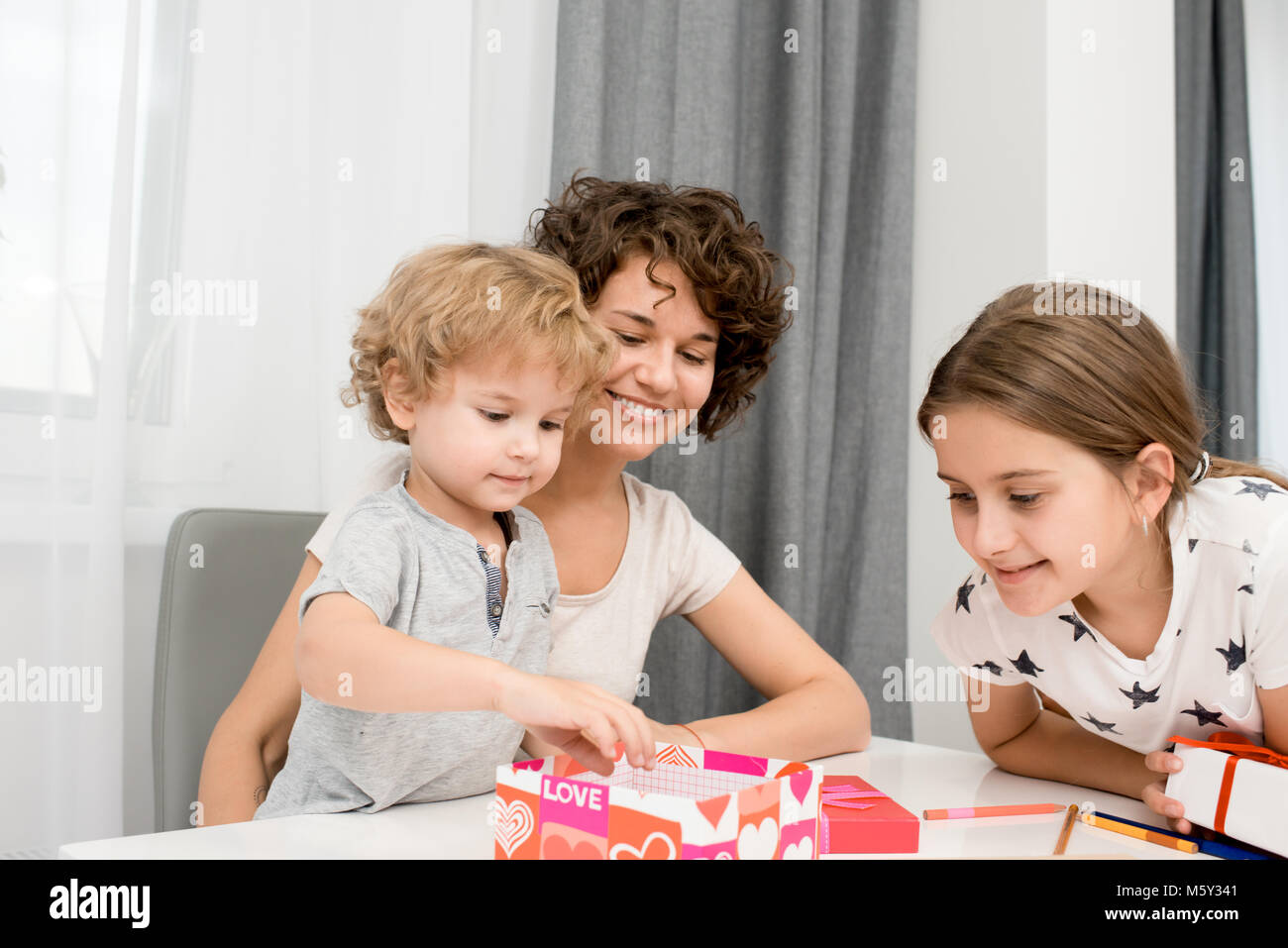 Portrait of happy young woman packing gifts with two children sitting at table at home and smiling Stock Photo