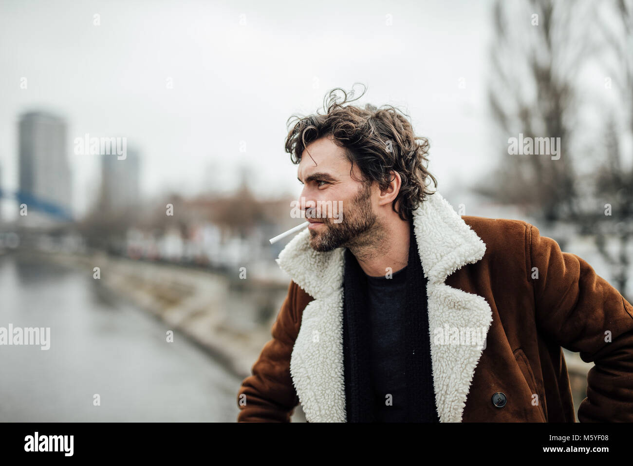 cool handsome man smoking cigarette outside in a aviator coat with masculine attitude waiting for some Stock Photo