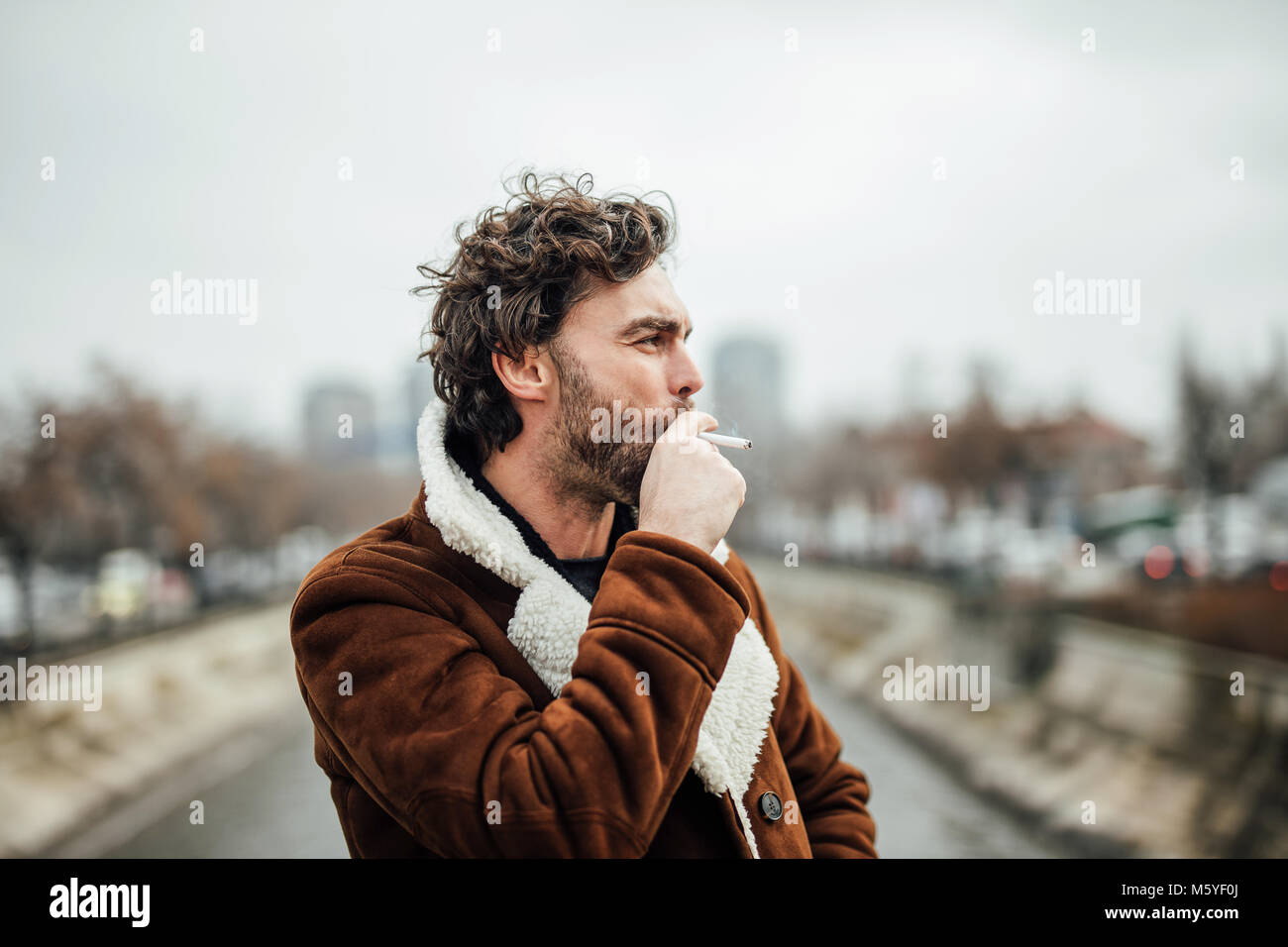 cool handsome man smoking cigarette outside in a aviator coat with masculine attitude waiting for some Stock Photo