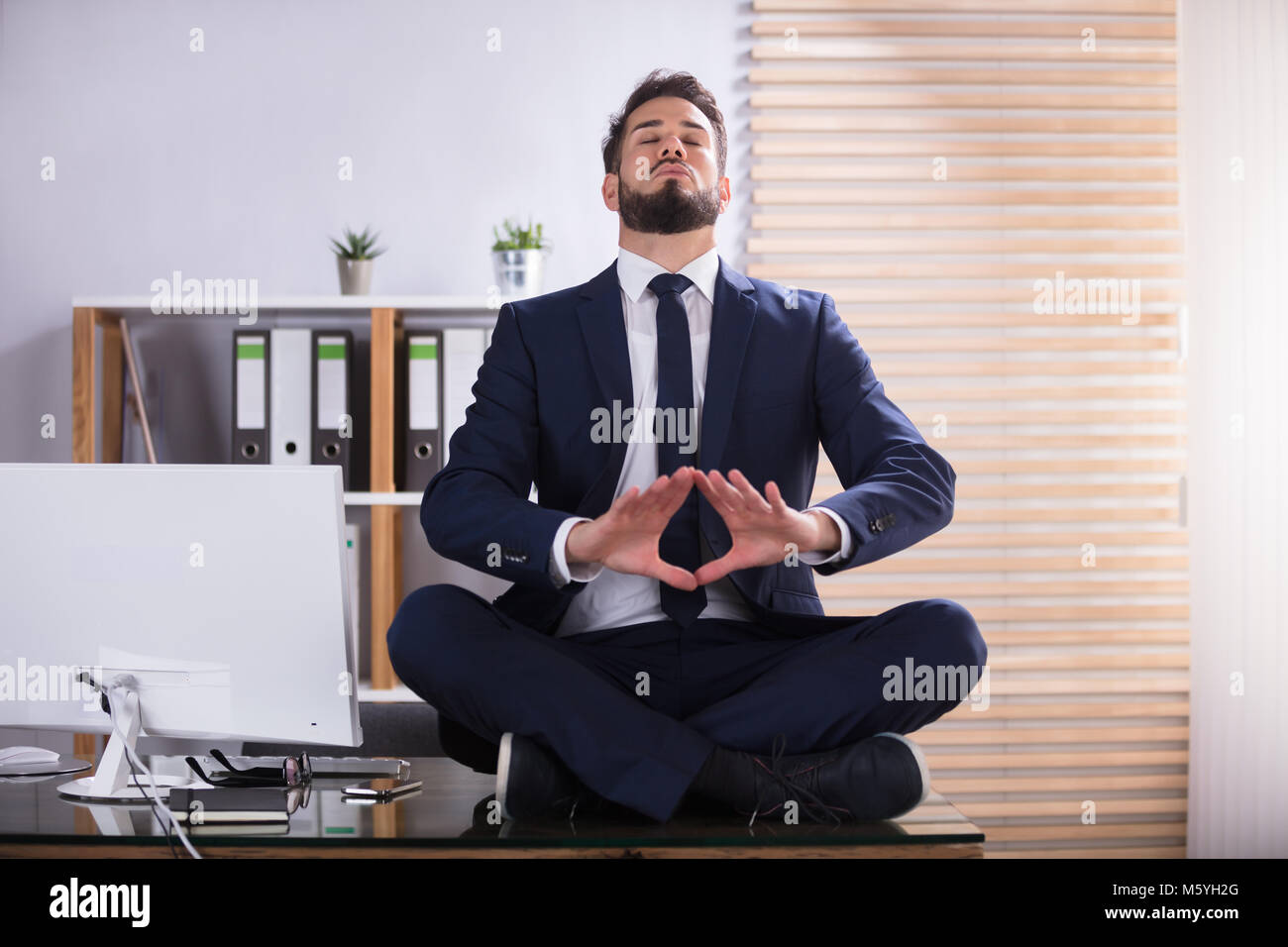 Young Businessman Sitting On Desk Doing Yoga In Office Stock Photo - Alamy