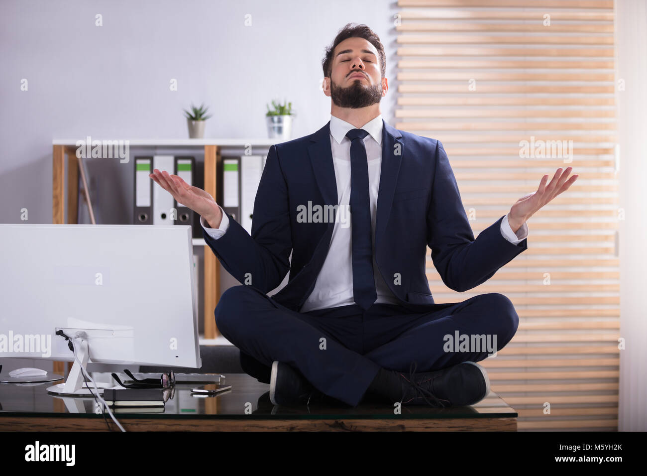 Young Businessman Sitting On Desk Doing Yoga In Office Stock Photo - Alamy