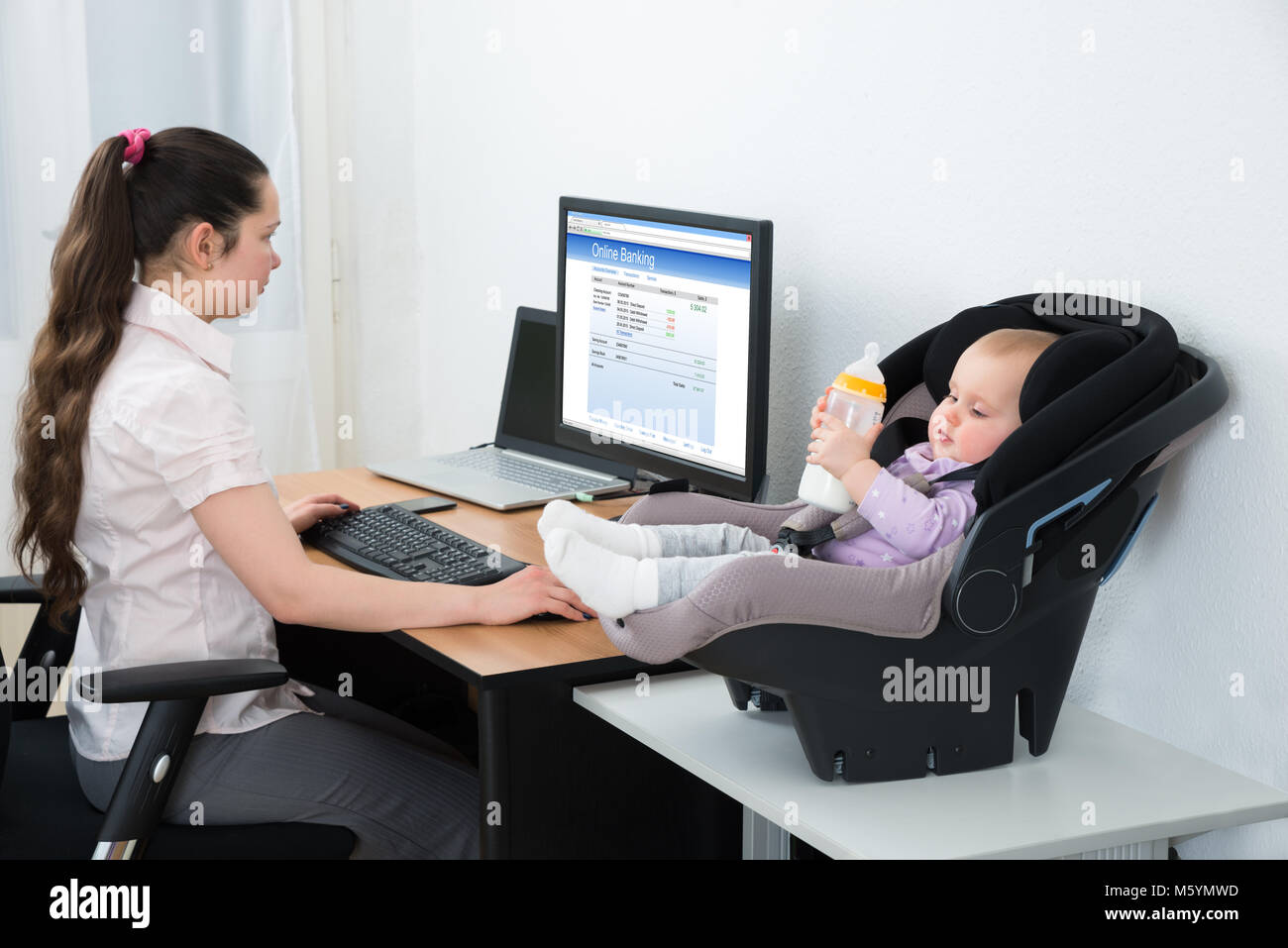 Woman Doing Online Banking On Computer With Baby Holding Milk Bottle Stock Photo