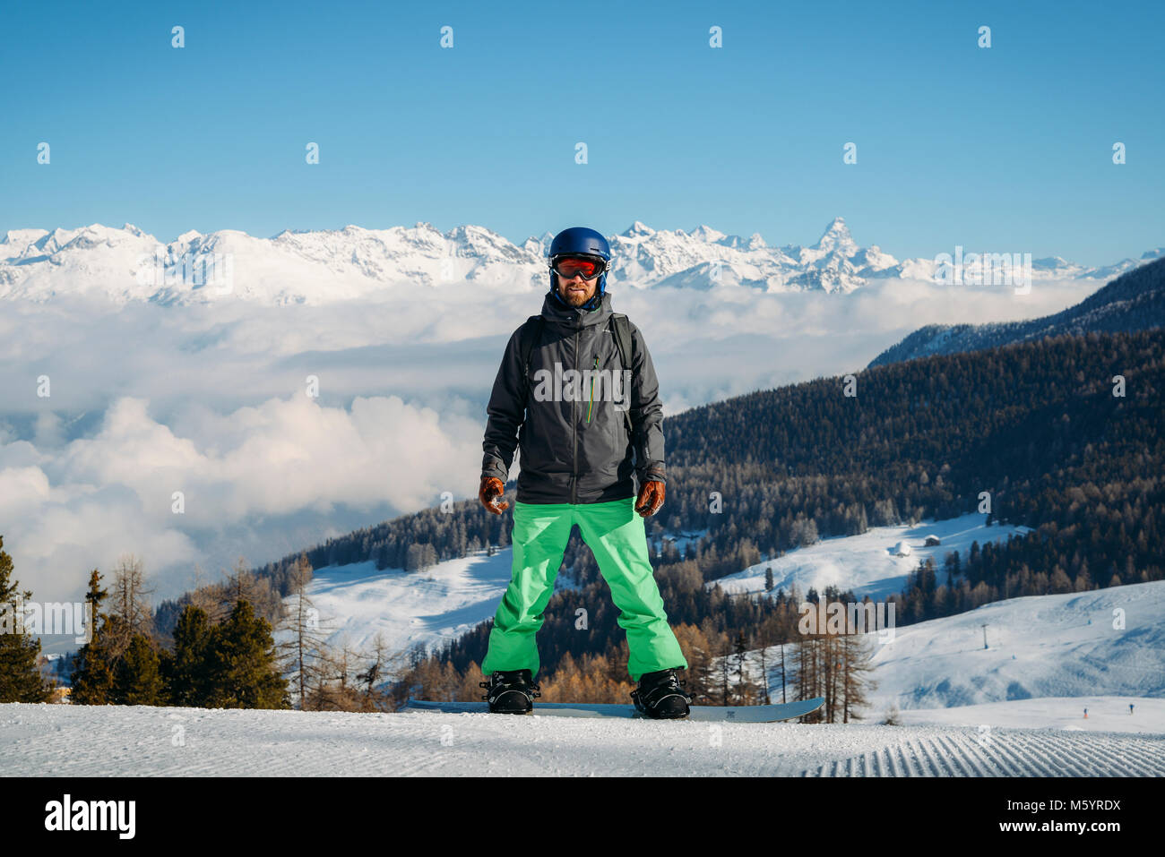 Full length shot of a smiling snowboarder standing on the slope in the mountains, looking to the camera at winter scene in Aosta, Italy Stock Photo