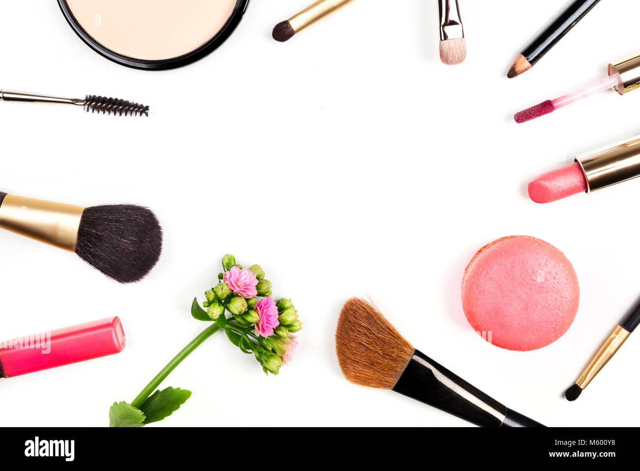 A frame of makeup brushes, powder, lipstick, and a mascara applicator, shot from above on a white background with a pink flower and a macaroon, with a Stock Photo