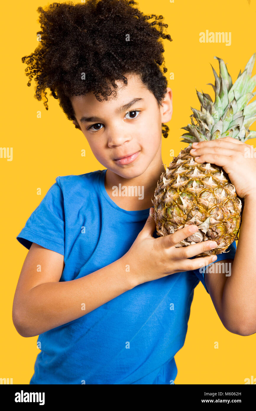 Happy little boy with a blue t-shirt holding a pineapple on his shoulder. Isolated on yellow Stock Photo