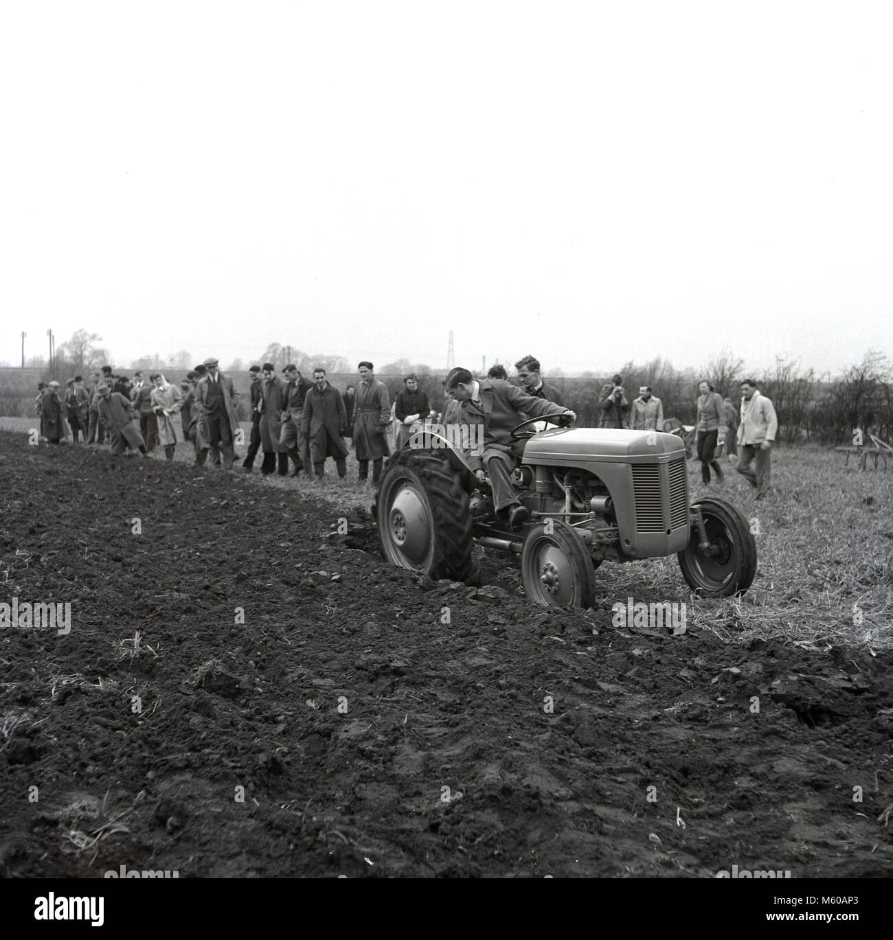 1950s, historical, standing in a line outside in a ploughed field, a group of agricultural students looking at a young male student driving a tractor and at the same time ploughing the field, England, UK. In the post war years in Britain with continued food rationing, there was a special emphasis put on training young people about agriculture, farming methods and food production. Stock Photo