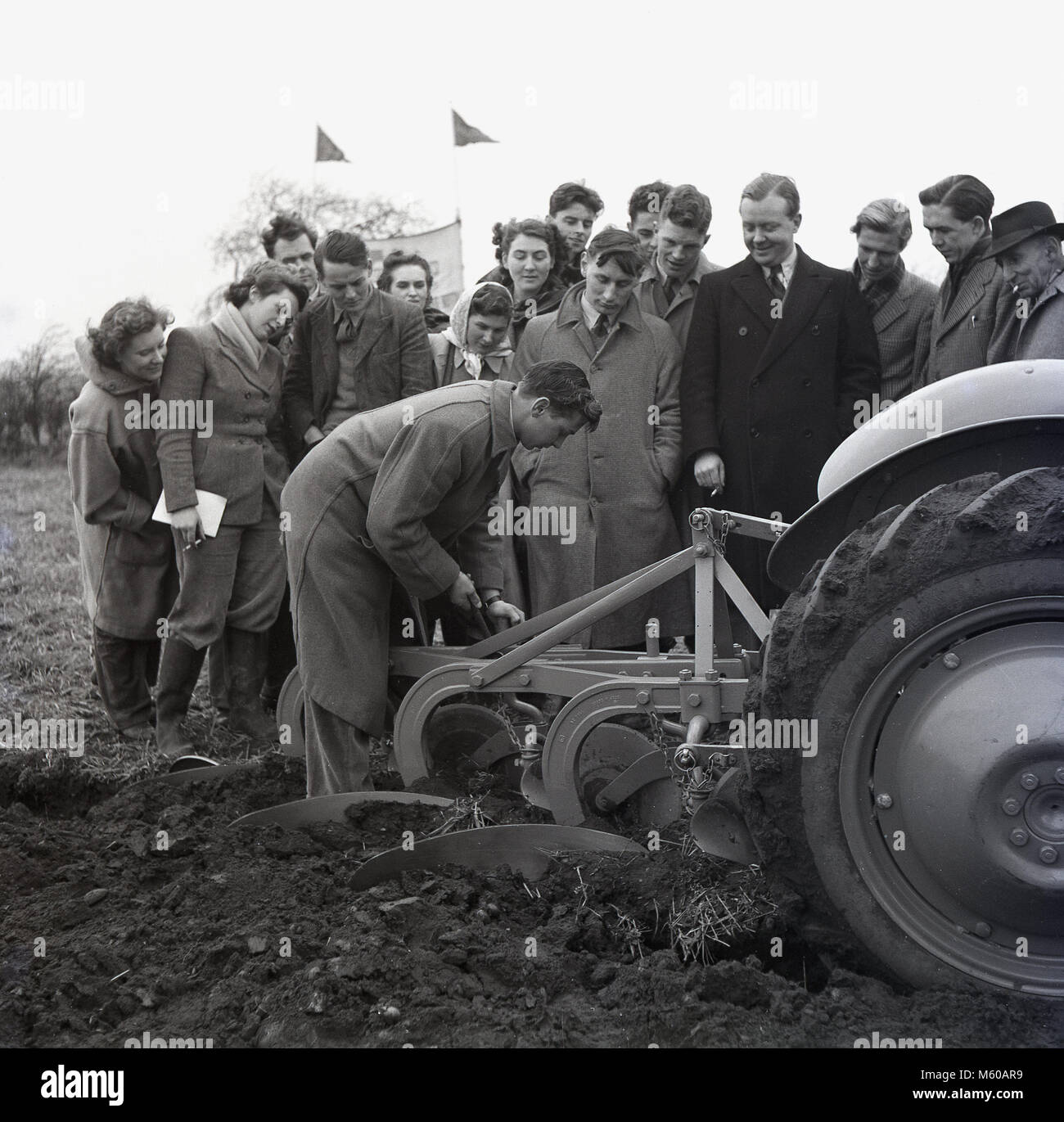 1950s, historical, standing outside in a ploughed field, a group of agricultural students looking at a young male student as he adjusts the levels of a metal plough attached to the back of a tractor, England, UK. In the post war years in Britain with continued food rationing, there was a special emphasis put on training young people about agriculture, farming methods and food production. Stock Photo