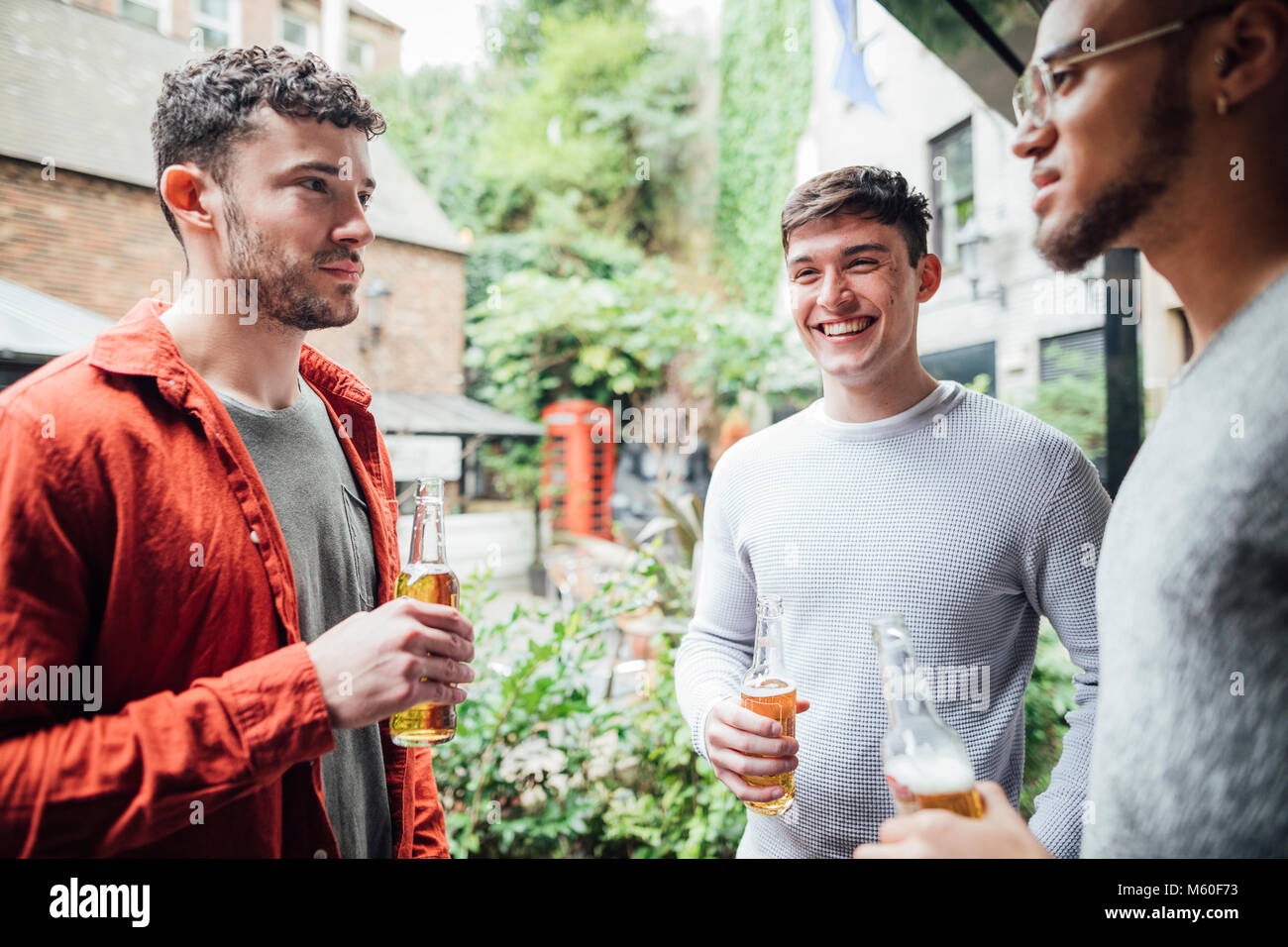 Three men are enjoying drinks together in the courtyard of a bar. They are talking and drinking bottles of beer. Stock Photo