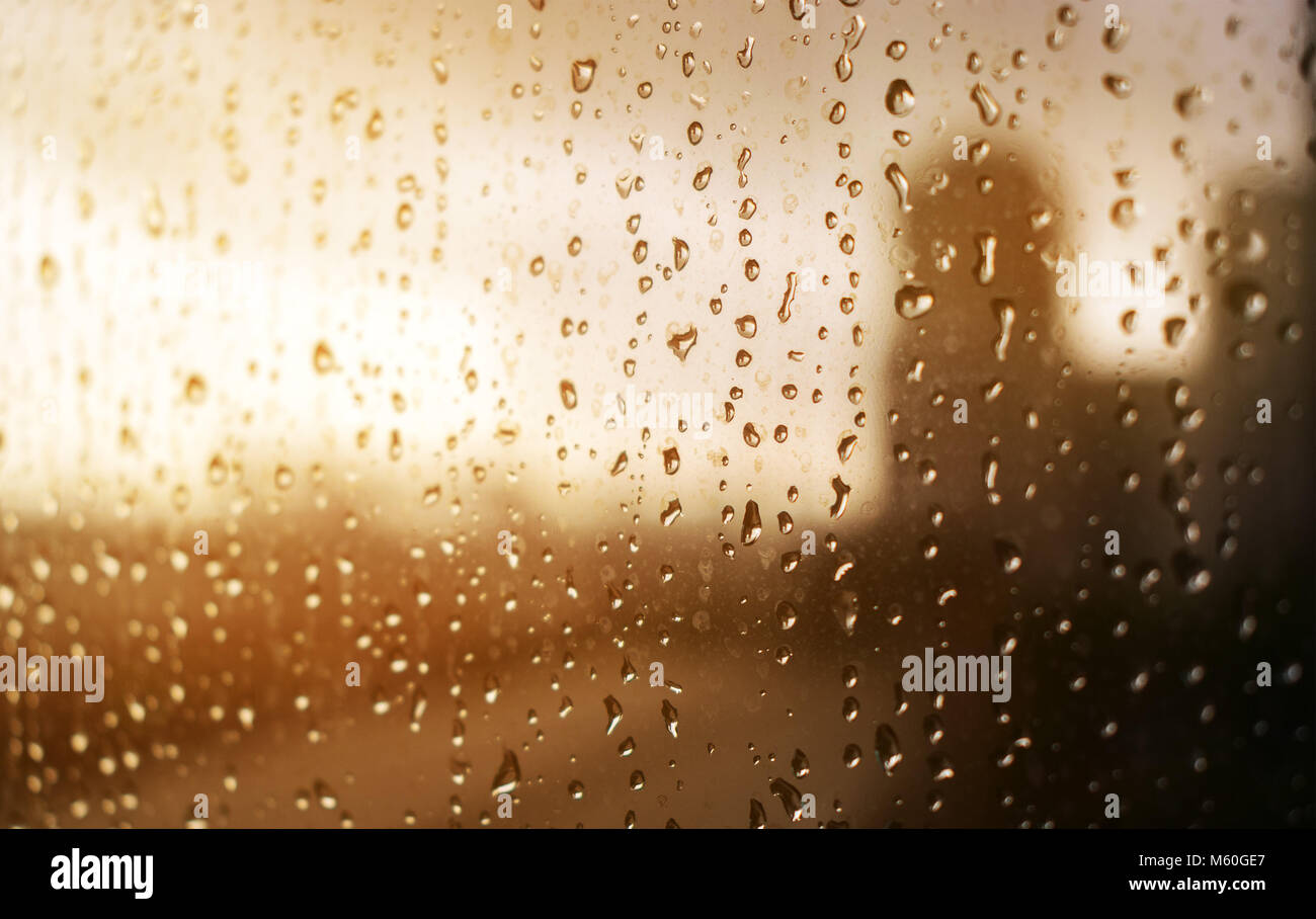 Water rain drops on a glass window, with a city in the background, warm yellow and orange color filter during sunset Stock Photo