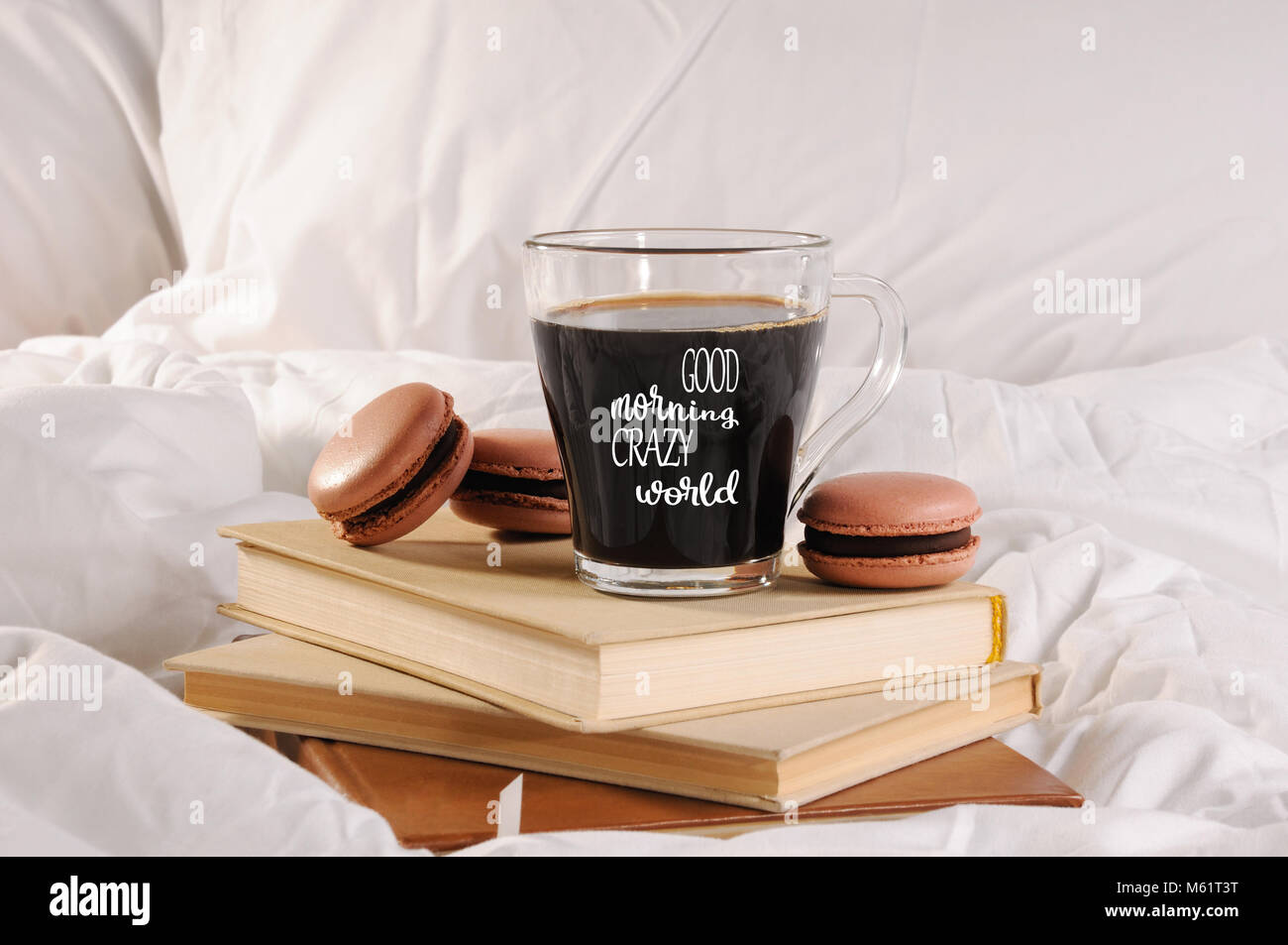 Morning cup of coffee with chocolate cakes Macaroons, on a pile of books in bed.  The inscription on the cup is  «Good morning crazy world». Stock Photo