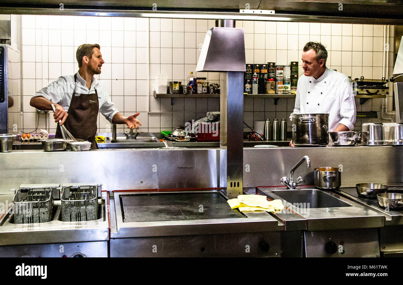 German two-Michelin-star chef Michael Kempf (left) talks to his colleague Christian Stockmann across the hob in the kitchen of the Parkhotel Ahrensburg, Germany. Both are wearing white chef’s jackets; spice jars and metal jugs are lined up on the shelf between them. Two deep-fat fryers with baskets hanging inside them can be seen in the foreground. Stock Photo