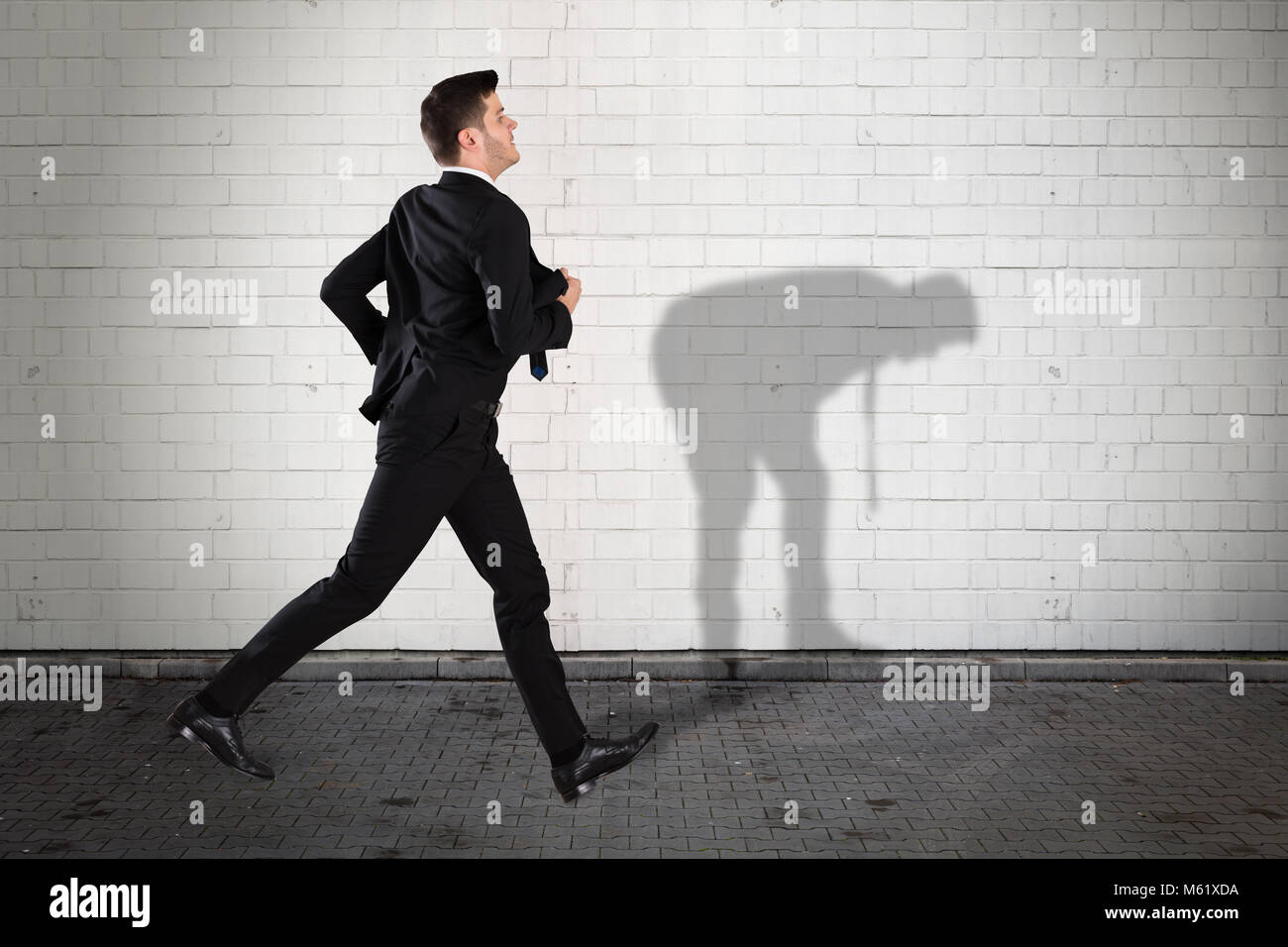 Young Businessman Running On Sidewalk With His Shadow Falling On Wall Stock Photo