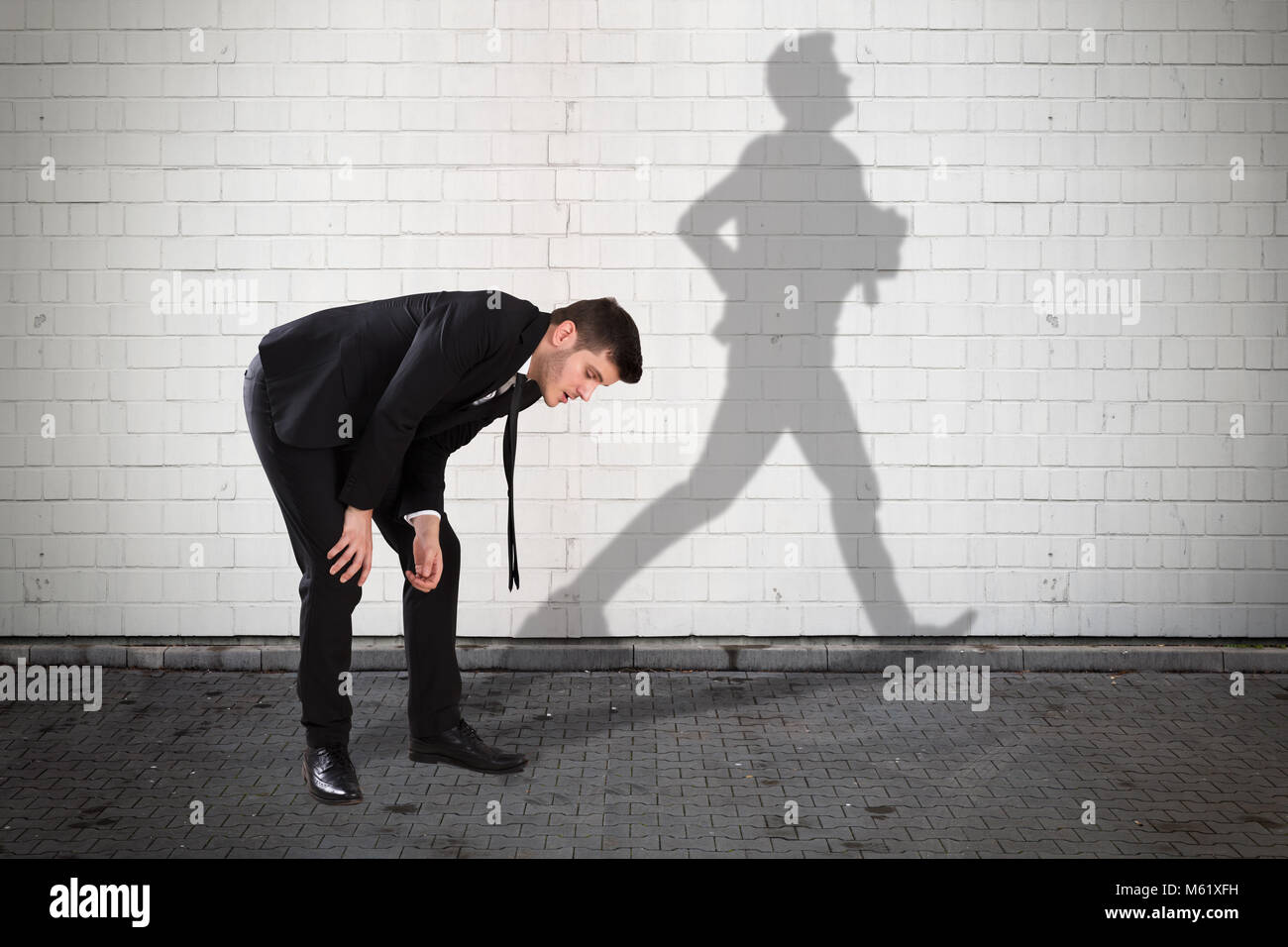 Young Tired Businessman With Shadow Of Man Running Formed On Wall Stock Photo