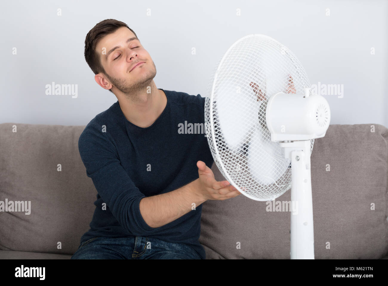 Young Man Sitting On Couch Enjoying Breeze With Electric Fan At Home Stock Photo