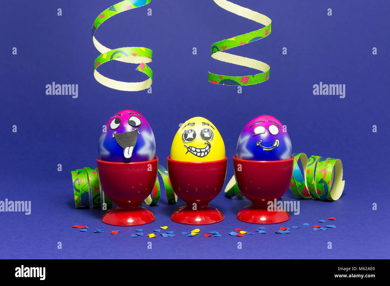 Group of colorful painted Easter eggs with funny cartoon style faces in red plastic egg cups and colorful confetti with paper streamers on purple back Stock Photo
