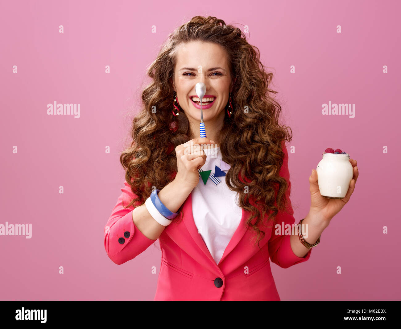 Pink Mood. happy modern woman with long wavy brunette hair isolated on pink background with farm organic yogurt and spoon having fun time Stock Photo