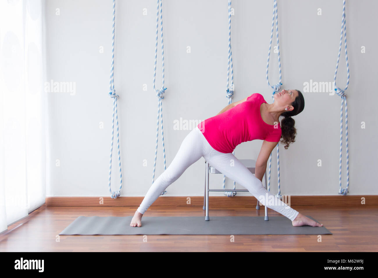 Young yoga instructor in utthita trikonasana pose with chair prop aid for torsion. Yoga teacher practices in studio. Wellness, healthy concept Stock Photo