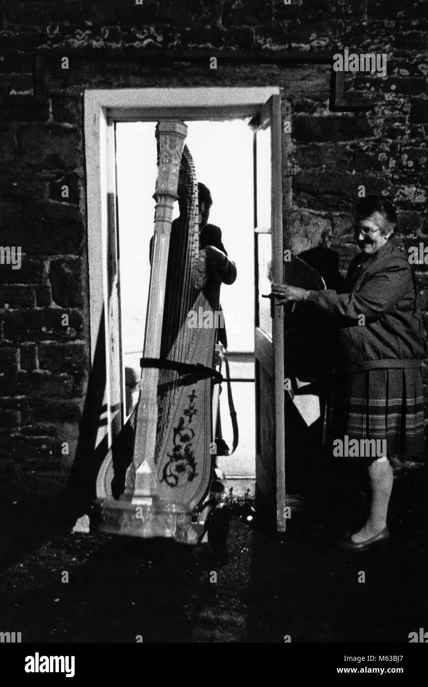 Man pushing large concert harp through door held open by elderly lady at small eisteddfod in village hall Talsarnau Gwynedd Wales UK Stock Photo