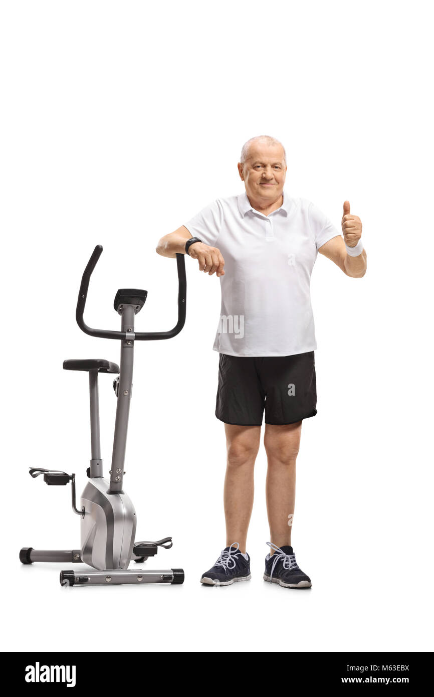 Full length portrait of a senior leaning on an exercise bike and making a thumb up gesture isolated on white background Stock Photo