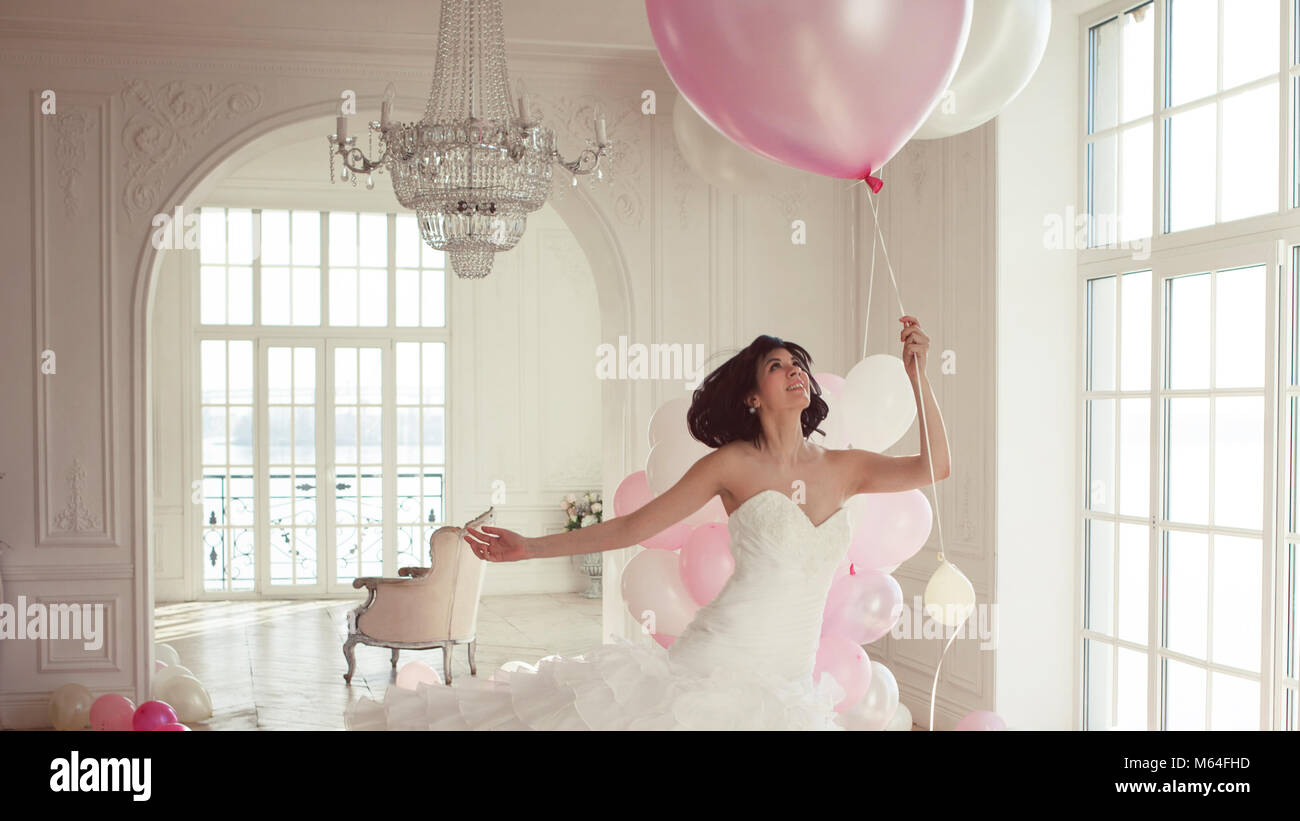 Young woman in wedding dress in luxury interior flies on pink and white balloons. Charming young bride brunette with short haircut in stylish Quincean Stock Photo