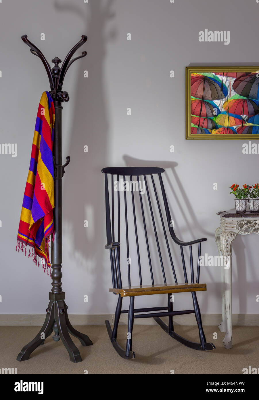 Interior shot of vintage rocking chair, coat hanger and flower pot on old style vintage table on background of off white wall with one hanged painting Stock Photo