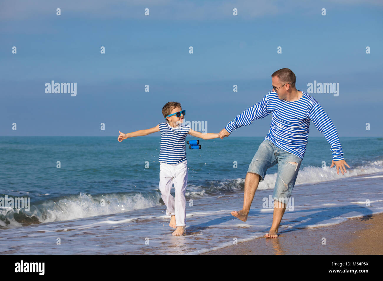 Father and son running on the beach at the day time. They are dressed in sailor's vests. Concept of sailors on vacation and friendly family. Stock Photo