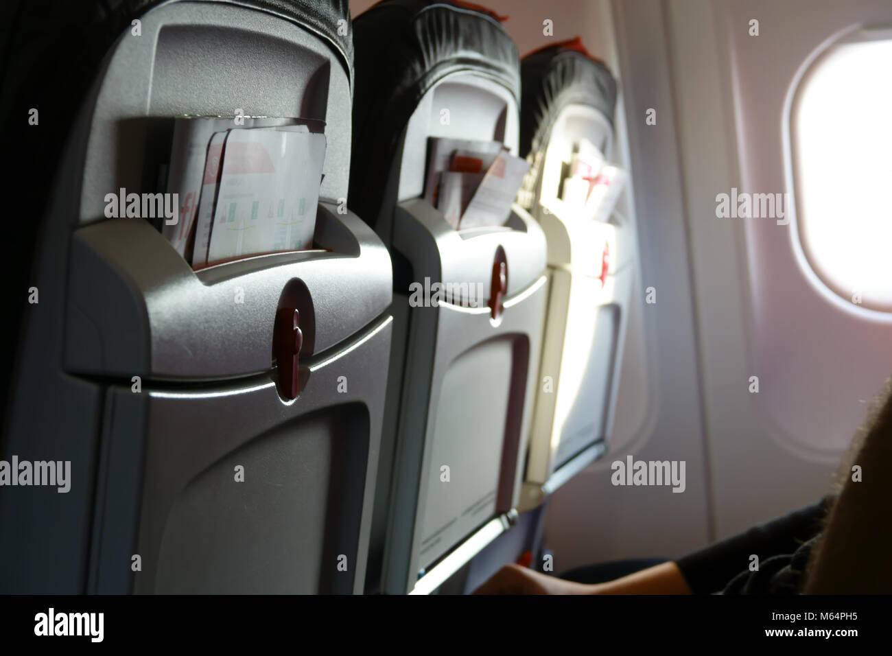 Three airplane seats with promotional brochures in aircraft passenger cabin. Light from porthole. People in chair. Stock Photo