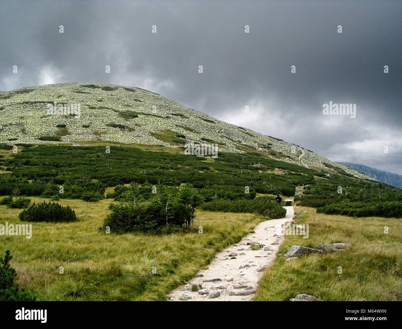 Stony hill with green vegetation and stony path under heavy rain clouds Stock Photo