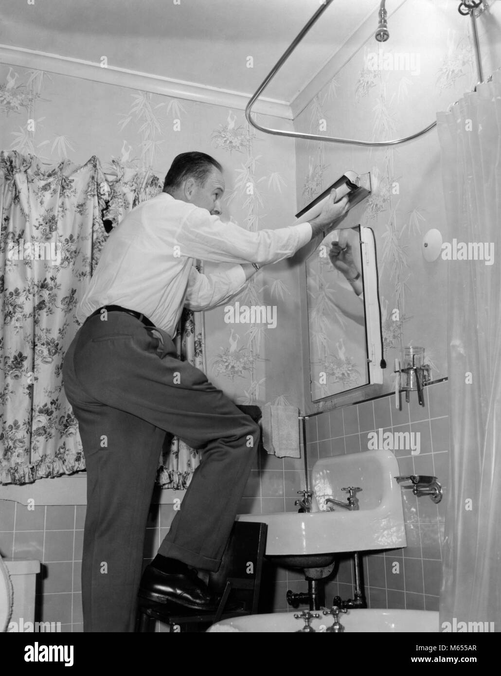 1950s MAN ON HOME REPAIR STEP STOOL IN BATHROOM REPLACING FLORESCENT LIGHTING FIXTURE ABOVE SINK - a1373 HAR001 HARS HOME IMPROVEMENT MALES MID-ADULT MID-ADULT MAN TASK AFTER WORK B&W BLACK AND WHITE CAUCASIAN ETHNICITY FIXTURE FLORESCENT HANDYMAN MISTER FIXIT OLD FASHIONED PERSONS REPLACEMENT REPLACING Stock Photo