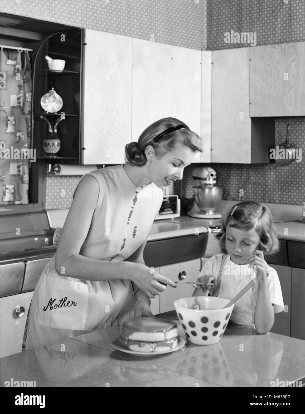 1950s MOTHER AND DAUGHTER IN KITCHEN FROSTING DOUBLE LAYER CAKE WORD MOTHER ON APRON - asp x16750 CAM001 HARS DAUGHTERS BOWLS INDOORS ICING NOSTALGIA HOMEMAKER HOMEMAKERS MOMS AND WORD LAYER FROSTING HOUSEWIVES MIXING BOWL JUVENILES B&W BLACK AND WHITE CAUCASIAN ETHNICITY KITCHEN ISLAND OLD FASHIONED PERSONS TEACHING SHOWING Stock Photo