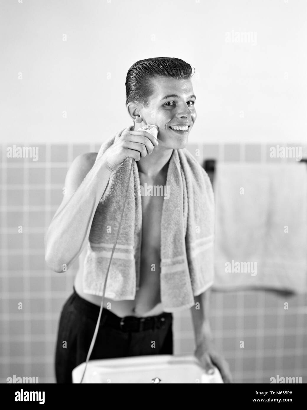 1950s SMILING TEENAGED BOY STANDING AT BATHROOM SINK SHAVING WITH ELECTRIC RAZOR LOOKING AT CAMERA - asp x16766 CAM001 HARS CONFIDENCE NOSTALGIA EYE CONTACT GROOMING HAPPINESS SHAVE TILE HYGIENE GROWTH SMILES TEENAGED JUVENILES MALES YOUNG ADULT MAN B&W BLACK AND WHITE CAUCASIAN ETHNICITY GOOD GROOMING LOOKING AT CAMERA LOOKING INTO MIRROR OLD FASHIONED PERSONS Stock Photo