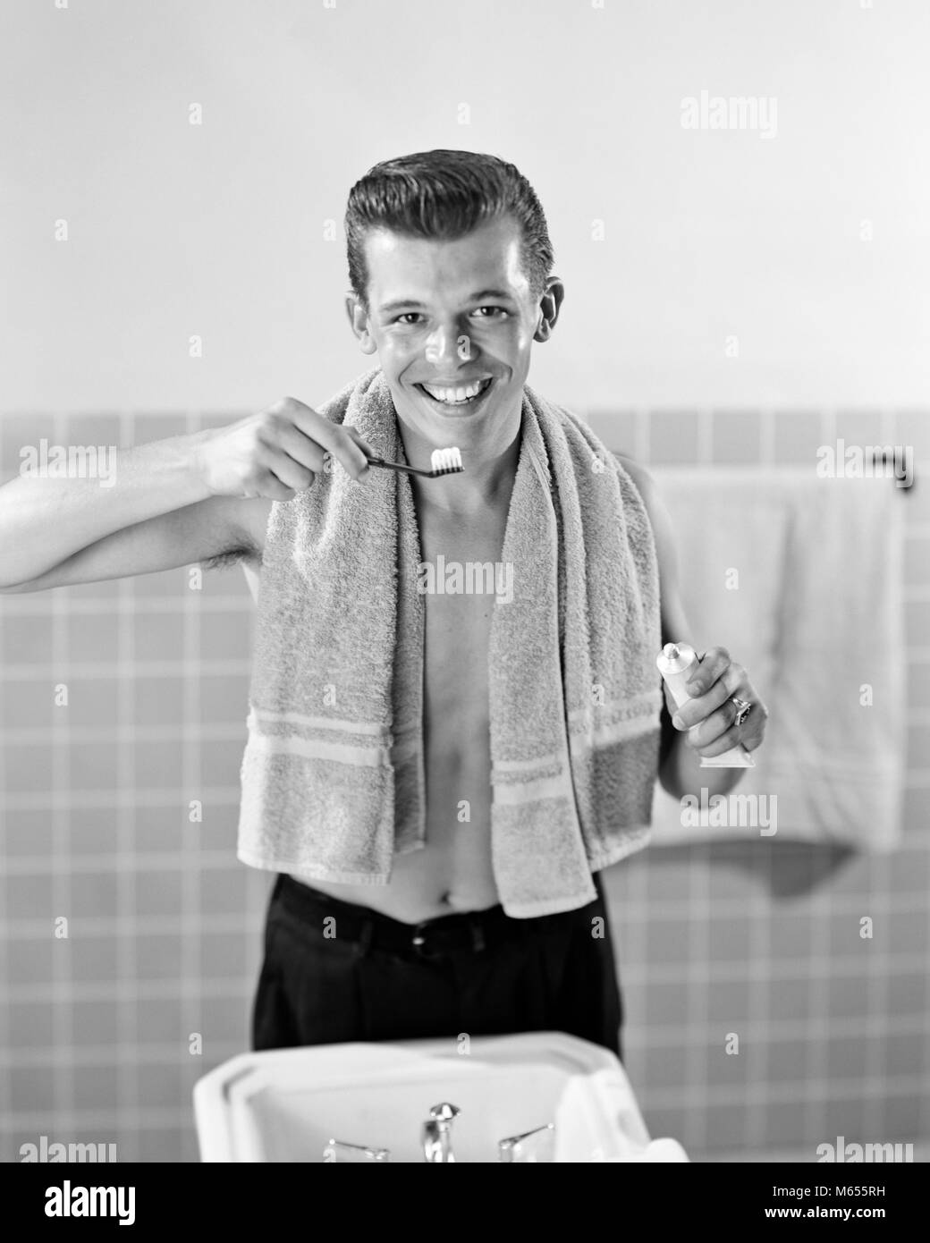 1950s SMILING TEENAGED BOY STANDING AT BATHROOM SINK BRUSHING TEETH LOOKING AT CAMERA - asp x16767 CAM001 HARS EYE CONTACT 16-17 YEARS GROOMING TOOTHBRUSH HAPPINESS TILE 18-19 YEARS HYGIENE GROWTH SMILES TEENAGED JUVENILES MALES YOUNG ADULT MAN B&W BLACK AND WHITE CAUCASIAN ETHNICITY DENTAL CARE DENTAL HEALTH GOOD GROOMING LOOKING AT CAMERA LOOKING INTO MIRROR OLD FASHIONED PERSONS TOOTHPASTE Stock Photo