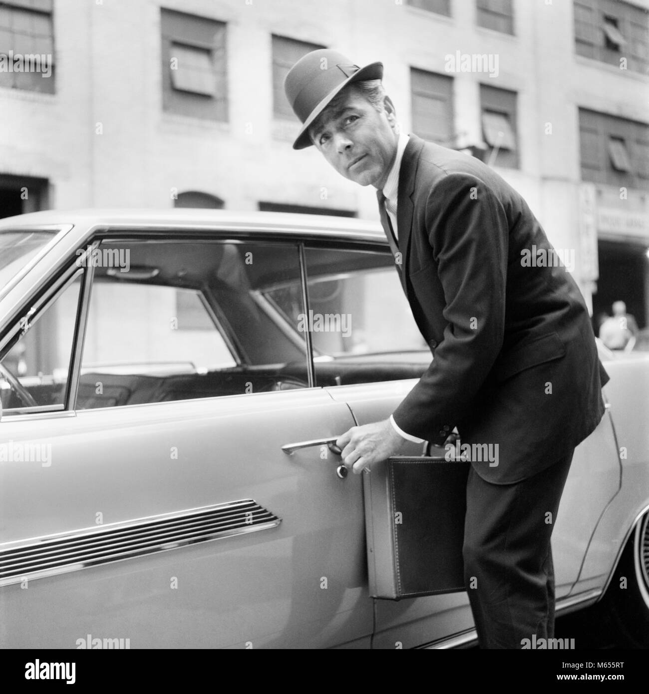 1960s BUSINESSMAN CARRYING BRIEFCASE UNLOCKING OPENING CAR DOOR LOOKING AT CAMERA - asp x19196 CAM001 HARS 35-40 YEARS 40-45 YEARS COMMUTE SUIT AND TIE SELLING AUTOS LOW ANGLE AUTOMOBILES VEHICLES SALESMEN MALES MID-ADULT MID-ADULT MAN UNLOCKING B&W BLACK AND WHITE CAUCASIAN ETHNICITY CLOSING COMMUTER LOCKING LOOKING AT CAMERA MAD MAN OCCUPATIONS OLD FASHIONED PERSONS Stock Photo