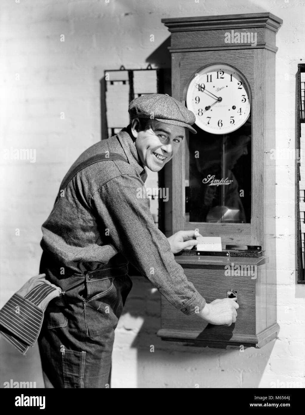 1930s SMILING MAN WORKER PUNCHING IN OUT AT TIME CLOCK LOOKING AT CAMERA - bx001776 CAM001 HARS OVERALLS INDOORS PROFESSION NOSTALGIA EYE CONTACT 25-30 YEARS 30-35 YEARS SKILL OCCUPATION HAPPINESS SKILLS CHEERFUL CAREERS HOURS VERTICAL PUNCHING SHIFT SMILES JOYFUL FRIENDLY EMPLOYEE ARRIVAL MALES MID-ADULT MID-ADULT MAN TIME CLOCK YOUNG ADULT MAN B&W BLACK AND WHITE DEPARTURE LOOKING AT CAMERA OCCUPATIONS OLD FASHIONED PERSONS TIME CARD Stock Photo