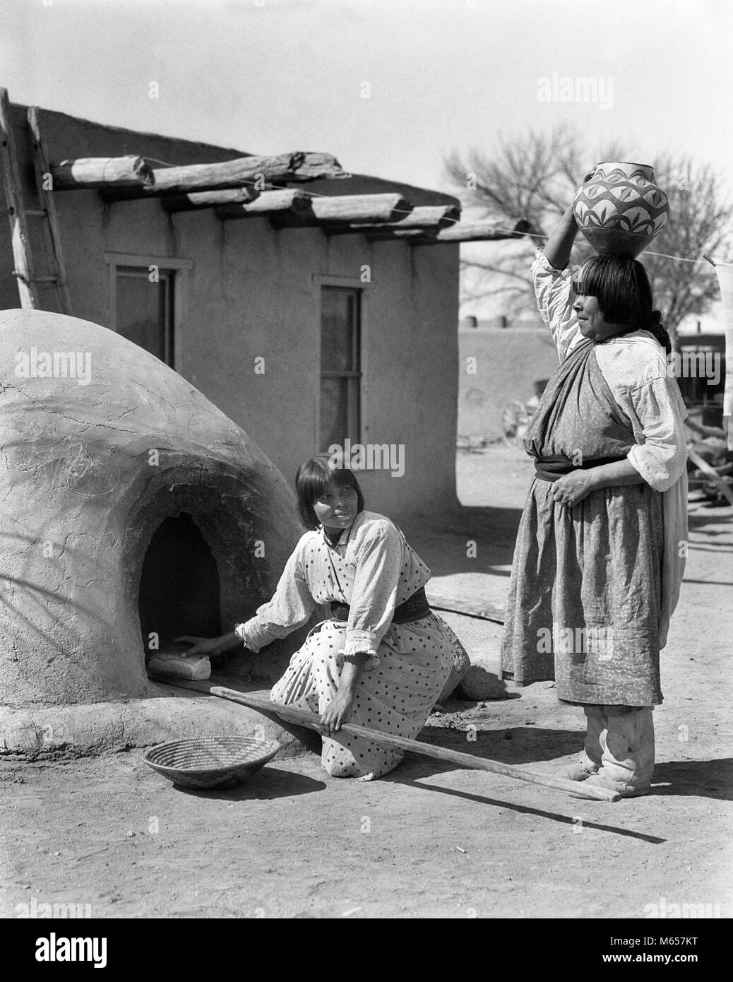 1930s TWO NATIVE AMERICAN WOMEN CARRYING OLLA WATER JAR ON HEAD AND BAKING BREAD IN HORNO OVEN SAN ILDEFONSO PUEBLO NM USA - i1559 HAR001 HARS FRIENDSHIP FULL-LENGTH LADIES INDIANS SPIRITUALITY AMERICANA NOSTALGIA MIDDLE-AGED NORTH AMERICA TOGETHERNESS JAR 25-30 YEARS 45-50 YEARS NORTH AMERICAN MIDDLE-AGED WOMAN AND MUD NATIVE AMERICAN PUEBLO SAN ILDEFONSO MID-ADULT MID-ADULT WOMAN NEW MEXICO YOUNG ADULT WOMAN ADOBE-BUILT B&W BLACK AND WHITE HORNO OVEN INDIGENOUS OLD FASHIONED OLLA OUTDOOR OVEN PERSONS SAN ILDEFONSO SOUTHWEST SOUTHWESTERN Stock Photo