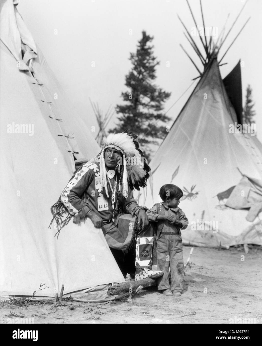1920s NATIVE AMERICAN INDIAN MAN CHIEF SITTING EAGLE AND BOY IN ENTRANCE TO TEPEE STONEY TRIBE OF BANFF ALBERTA CANADA - i525 HAR001 HARS RURAL HOME LIFE EAGLE COPY SPACE FRIENDSHIP HALF-LENGTH VILLAGE INDIANS NOSTALGIA FATHERS MIDDLE-AGED NORTH AMERICA TOGETHERNESS MIDDLE-AGED MAN 3-4 YEARS 45-50 YEARS NORTH AMERICAN TEEPEE AND TIPI CANADIAN CHIEF DADS LEADERSHIP PRIDE GROWTH CONNECTION GRANDCHILD GRANDFATHERS TEPEE STONEY TRIBE GRANDSON NATIVE AMERICAN ALBERTA CONICAL JUVENILES MALES NATIVE AMERICANS WESTERN CANADA B&W BANFF BLACK AND WHITE GRANDPA INDIGENOUS OLD FASHIONED PERSONS Stock Photo