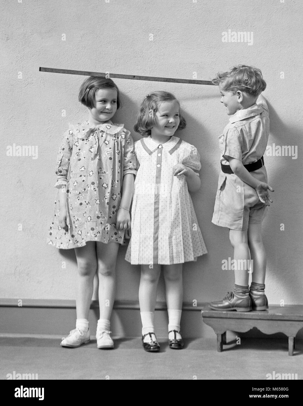 1930s BOY STANDING ON A STOOL MEASURING THE HEIGHT OF TWO GIRLS WITH YARD STICK - j2244 HAR001 HARS PLEASED JOY LIFESTYLE CELEBRATION FEMALES BROTHERS STOOL HEALTHINESS HOME LIFE COPY SPACE FRIENDSHIP FULL-LENGTH PHYSICAL FITNESS CARING INDOORS SIBLINGS SISTERS NOSTALGIA TOGETHERNESS 3-4 YEARS DRESSES GOALS SUCCESS 5-6 YEARS HAPPINESS CHEERFUL DISCOVERY STRATEGY EXCITEMENT PRIDE AUTHORITY GROWTH SIBLING SMILES CONNECTION JOYFUL COOPERATION SHORT PANTS SMALL GROUP OF PEOPLE SOLUTIONS CHILDHOOD JUVENILES MALES MARY JANE SHOES PRECISION YARD STICK B&W BLACK AND WHITE CAUCASIAN ETHNICITY COMPARING Stock Photo