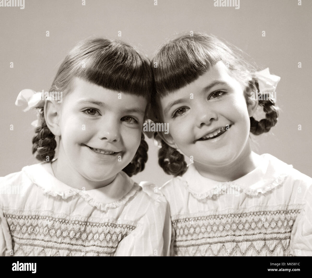 1940s 1950s SMILING TWIN GIRLS WITH PIGTAILS HEADS TOGETHER LOOKING AT CAMERA SMILING - j57 HAR001 HARS SISTERS NOSTALGIA TOGETHERNESS BRUNETTE MATCHING SAME HAPPINESS DIFFERENCE GROWTH SIBLING SMILES CONNECTION JOYFUL COOPERATION LOOK-ALIKE BRAIDS DUPLICATE JUVENILES LOOK ALIKE PIGTAIL PIGTAILS B&W BANGS BIGGER BLACK AND WHITE CAUCASIAN ETHNICITY CLONE HEAD TO HEAD LOOKING AT CAMERA OLD FASHIONED SAMENESS SMALLER SMOCKING Stock Photo