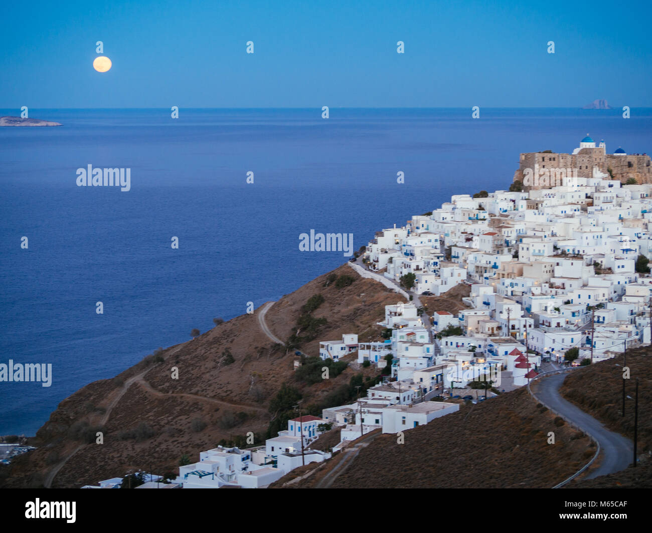 A lithe road leading to Chora of Astypalaia in blue hour and the full moon rising from the horizon Stock Photo