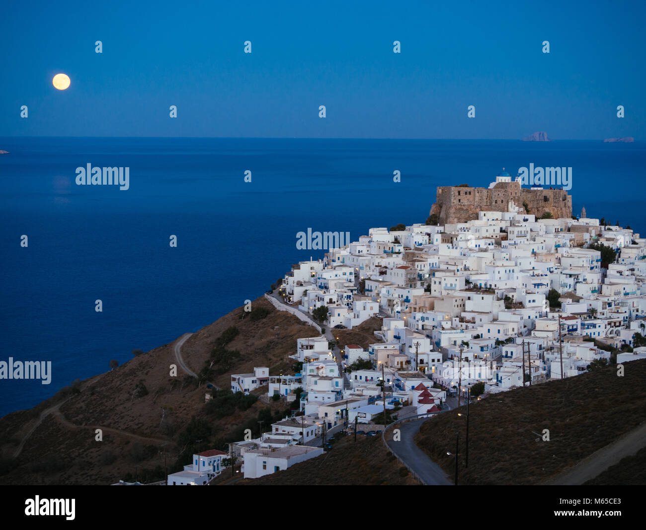 A lithe road leading to Chora of Astypalaia in blue hour and the full moon rising from the horizon Stock Photo