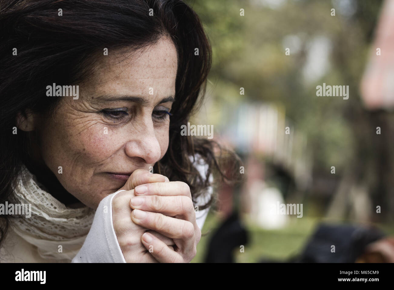 Portrait of mature woman concerned with hands together looking down in the park. Middle aged lady with pessimistic look. Sad, melancholic concept Stock Photo