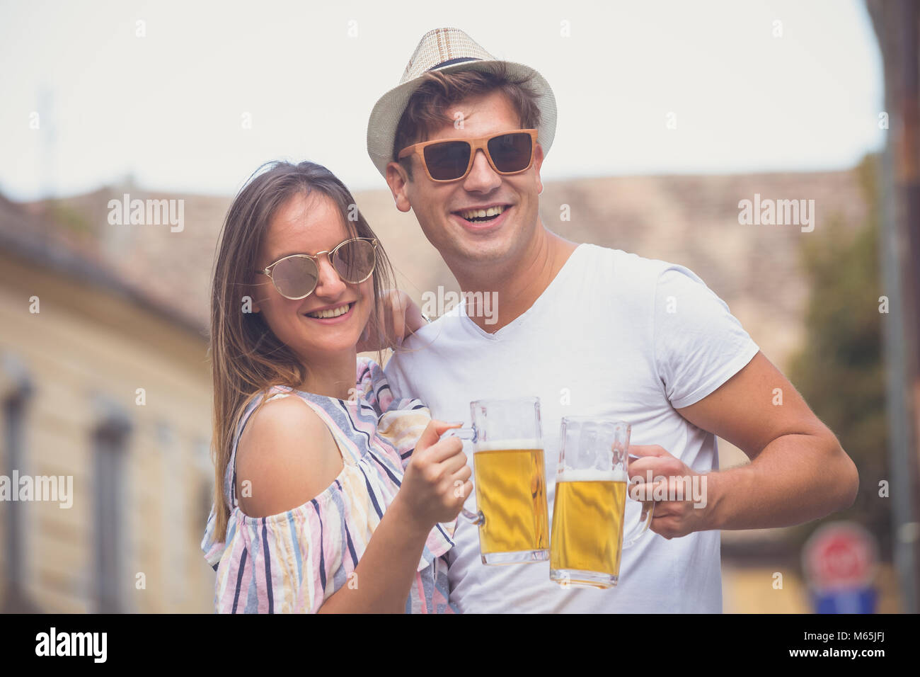 Young hipster couple in love cheering with beer mugs at the city street