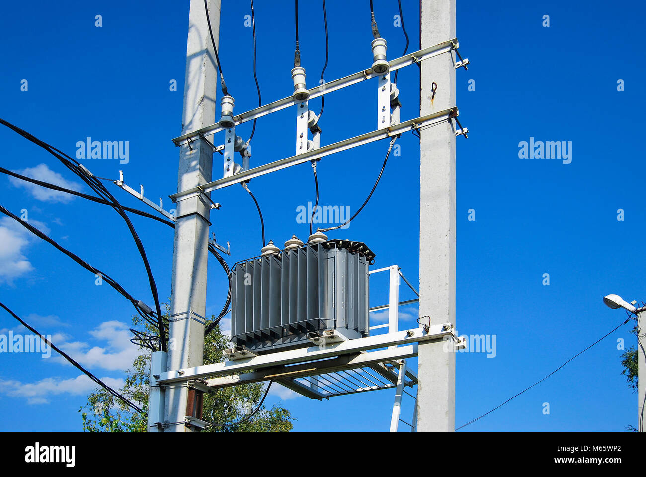 An electrical distribution transformer with cooling fins is located on the pole. Against the blue sky. Stock Photo