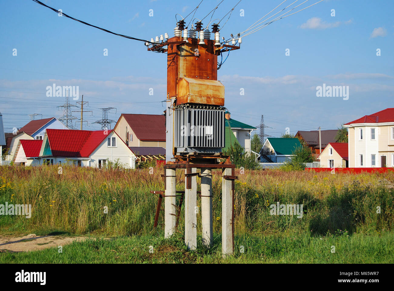Old rusty electrical distribution transformer with cooling fins in the village. Against the backdrop of village houses. Stock Photo