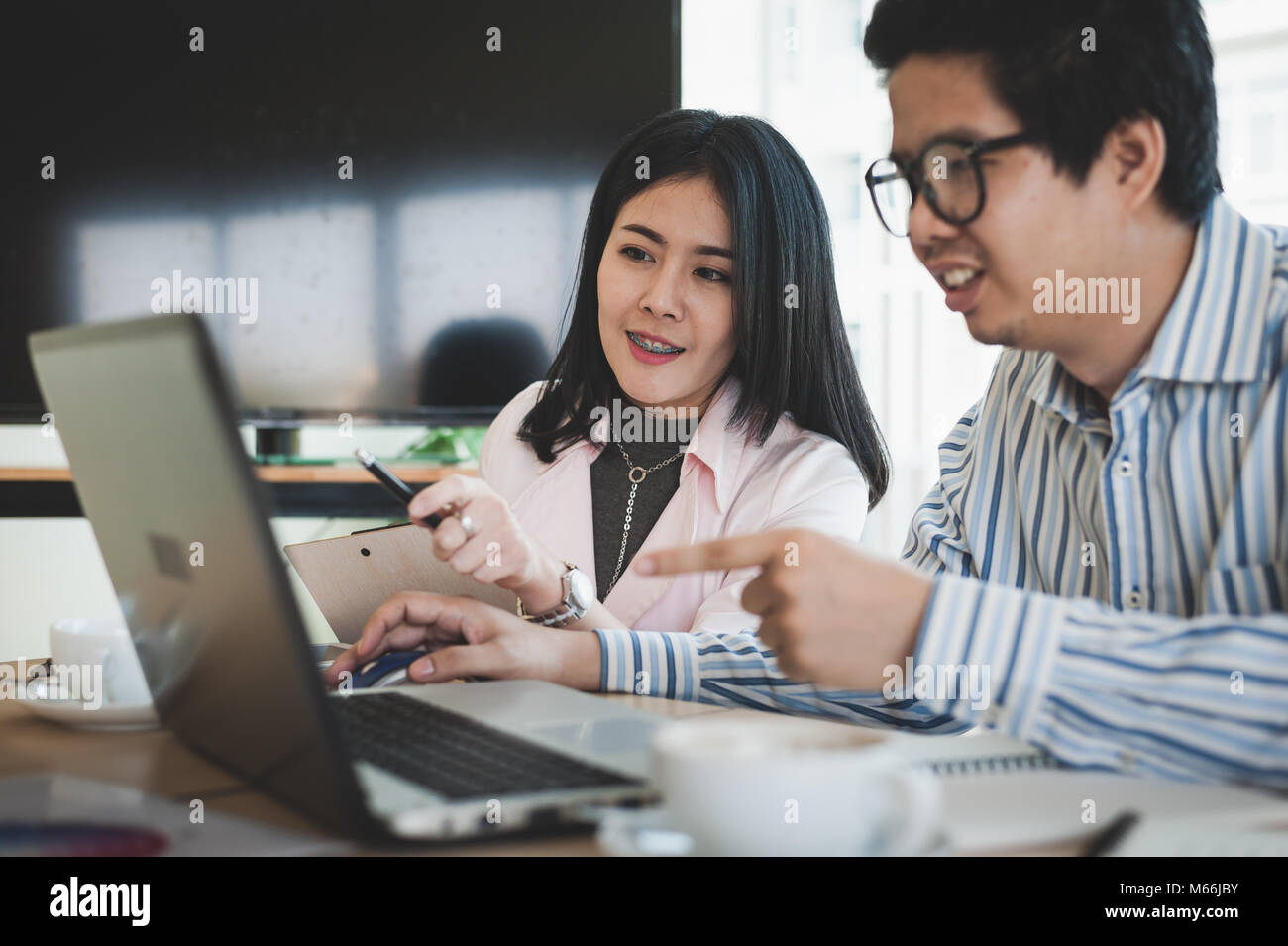 Young Asian businessman and businesswoman talking about their work while looking at laptop monitor in meeting room. Startup business teamwork concept. Stock Photo