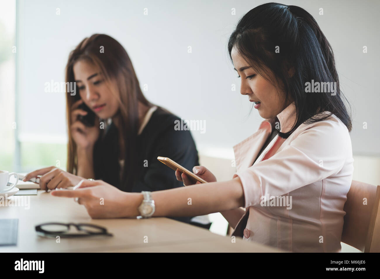Young Asian businesswoman talking on smartphone while sitting beside colleague on break time in office. Wireless communication concept Stock Photo